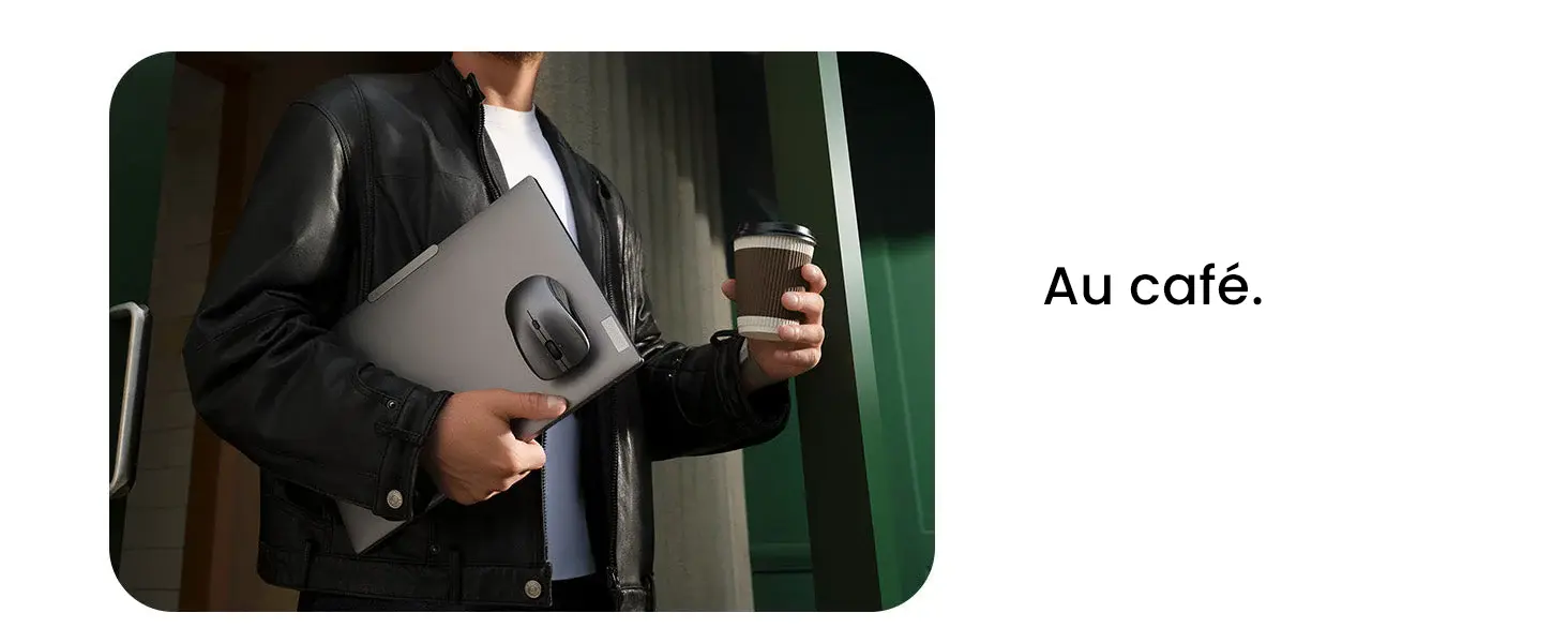 Une personne en veste de cuir noir et t-shirt blanc, sortant d'un bâtiment moderne, tient un ordinateur portable fermé avec la souris magnétique noire attachée et un gobelet de café à emporter.