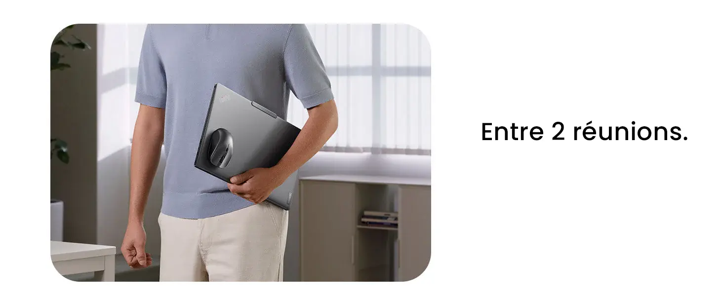 Une personne en polo bleu clair marche dans un bureau bien éclairé, tenant un ordinateur portable fermé sous le bras, avec la souris magnétique noire attachée au dos de l'appareil.