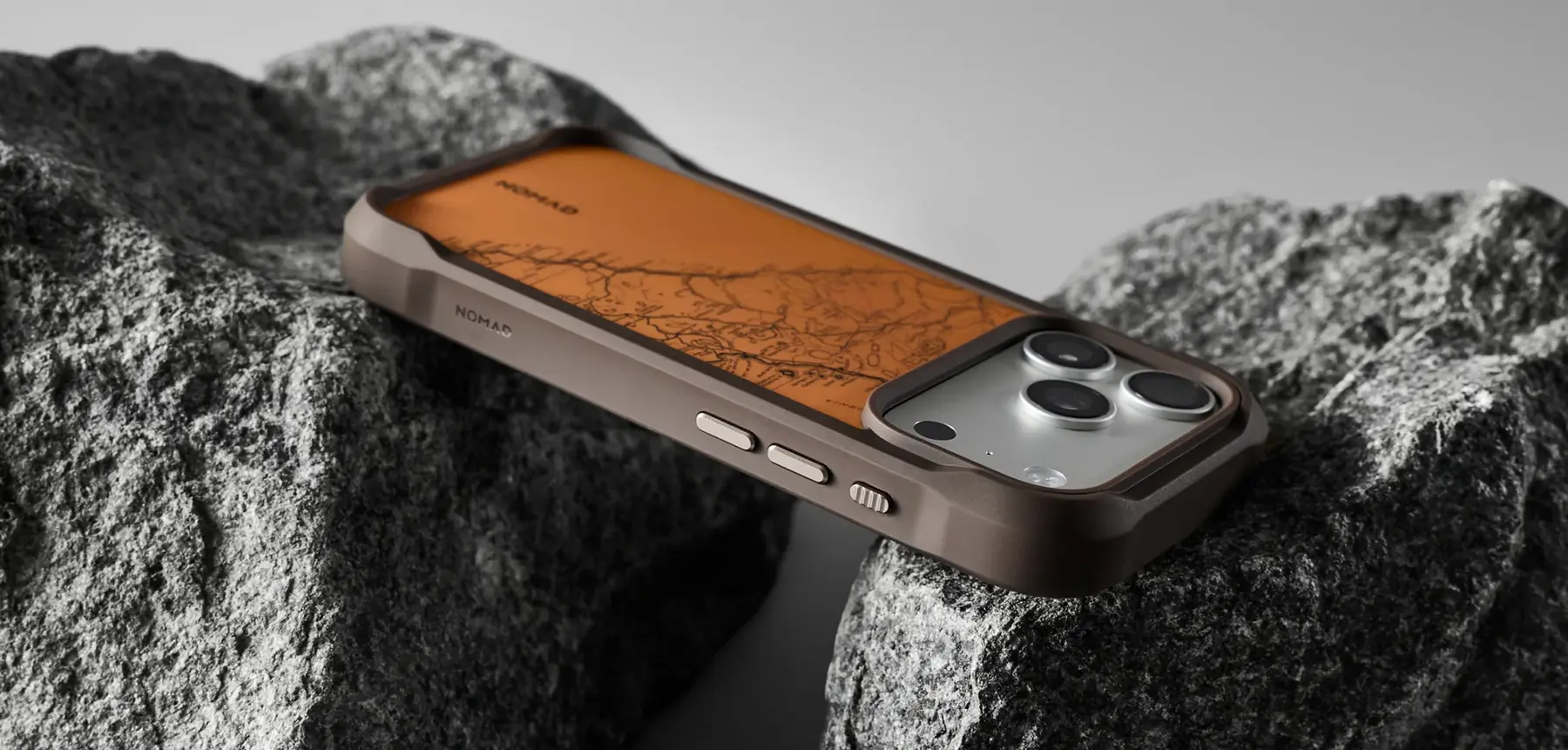 Coque MagSafe NOMAD Rugged Case Arroyo marron sur iPhone Pro, dos orange à motif cartographique, posée sur des rochers
