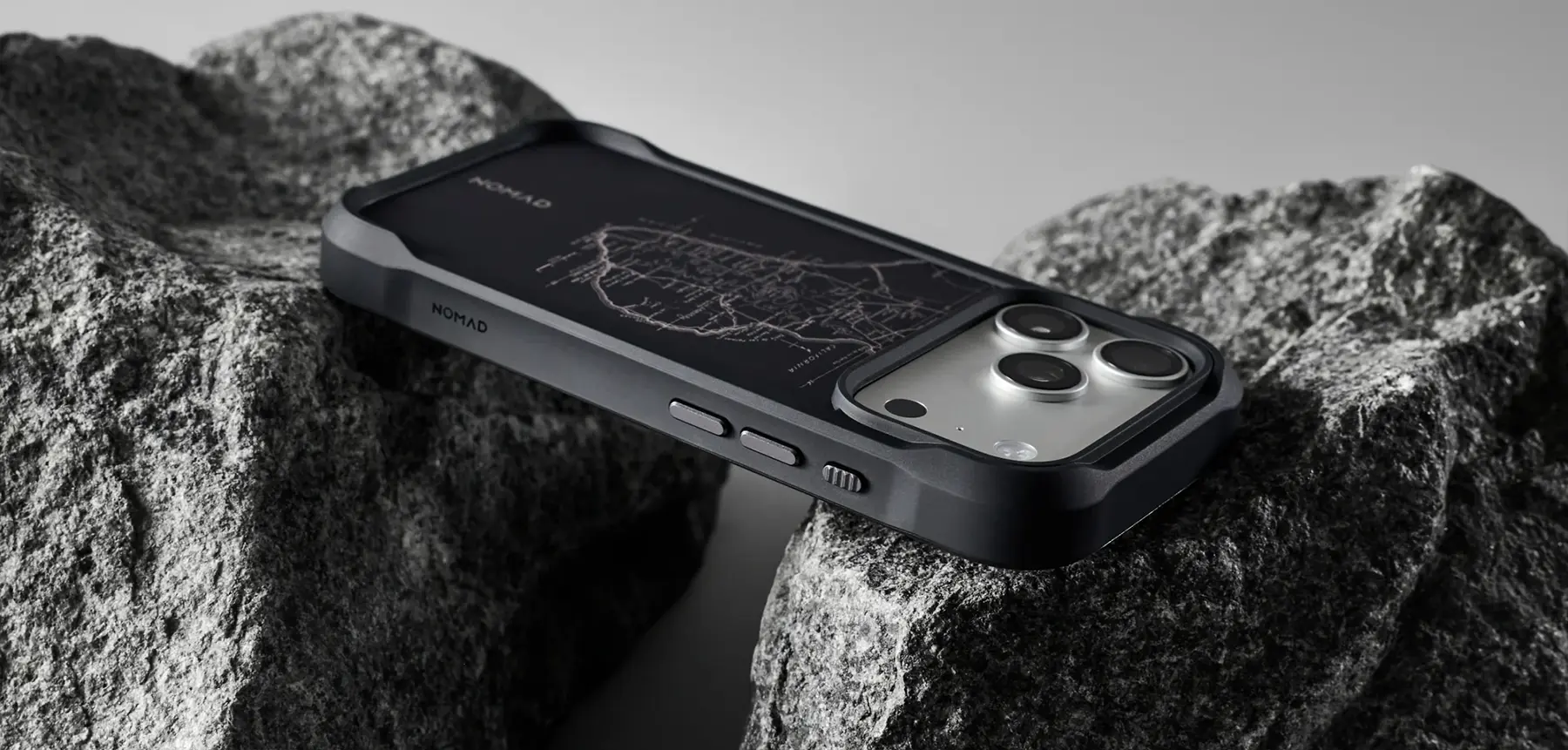 Coque MagSafe NOMAD Rugged Case Norte noir sur iPhone Pro, dos noir à motif cartographique, posée sur des rochers